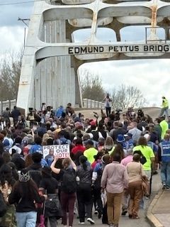 Crowd marching across Edmund Pettus Bridge during a protest, with signs and raised fists.