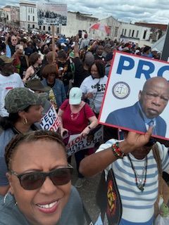 Crowd at protest holding signs, including one with a man’s portrait, in front of government buildings