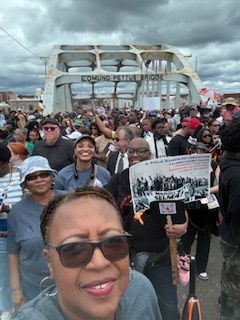 Crowd of people on a bridge march holding signs, with a smiling woman in sunglasses in the foreground.