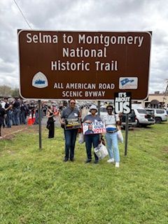 People posing by a Selma to Montgomery National Historic Trail sign on a grassy roadside, with a crowd behind them