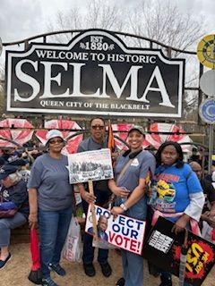 Four protesters holding signs in front of the historic Selma welcome sign, including “Protect Our Vote!”