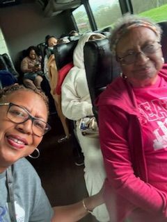 Two people smiling on a bus, one taking a selfie, with passengers and seats in the background.