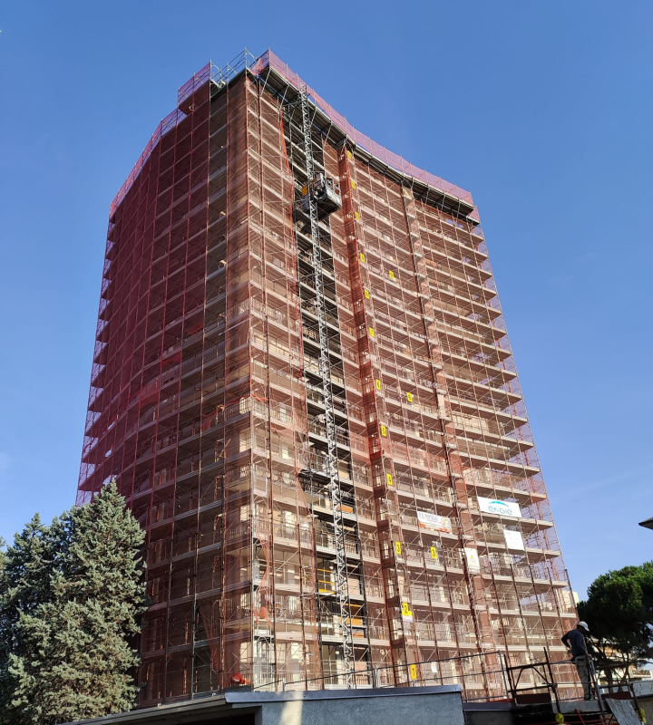 Un grattacielo imponente avvolto da impalcature arancioni si staglia contro un cielo azzurro.