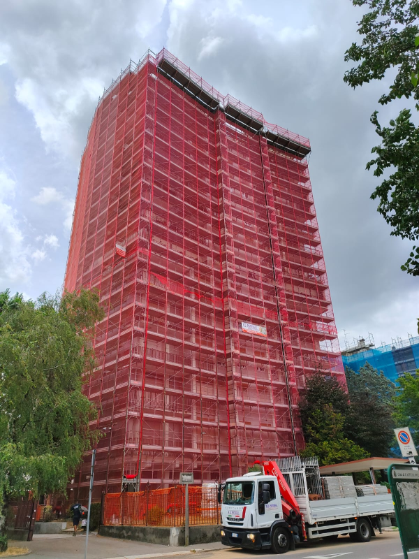 Un alto edificio avvolto da impalcature rosse con un camion bianco parcheggiato sotto, in una strada nuvolosa.