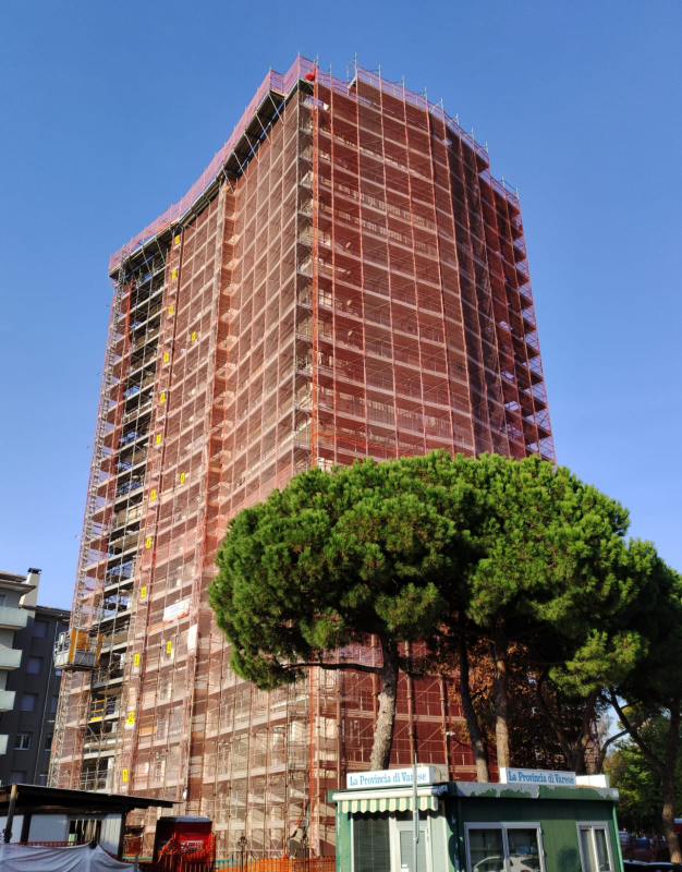 Un grattacielo in costruzione avvolto da impalcature rosse, con un grande albero verde di fronte.