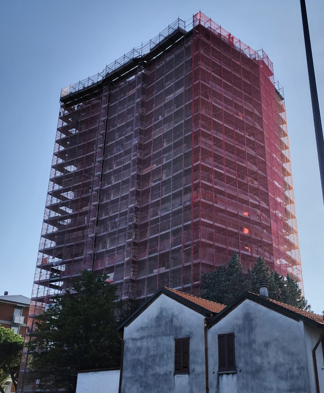 Un alto edificio in costruzione, avvolto da impalcature rosse, si staglia dietro due piccole case al crepuscolo.