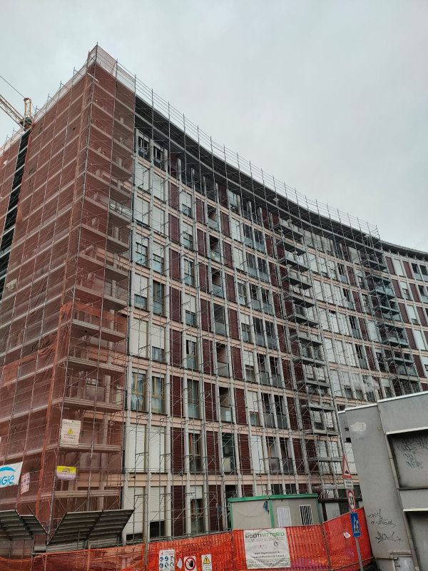 Edificio curvo a più piani in costruzione, con impalcature rosse e cantiere recintato.