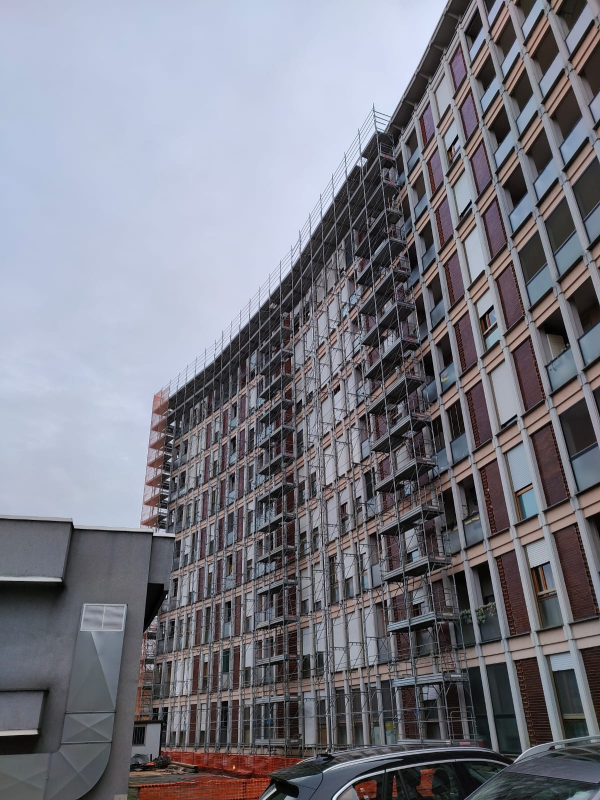 In una giornata nuvolosa, un moderno edificio residenziale dalle forme sinuose, ancora in costruzione, si affaccia su un parcheggio insieme ad alcune auto.