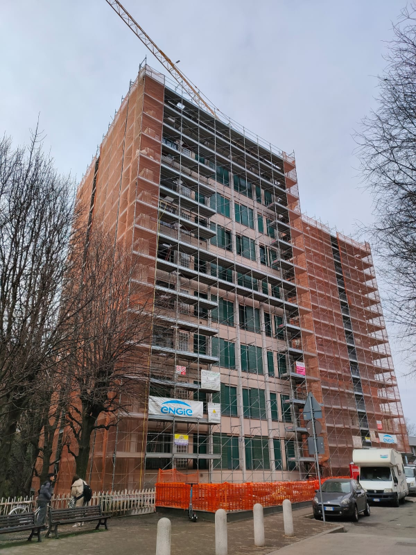 Alto palazzo residenziale in mattoni in costruzione, con impalcature, recinzioni di sicurezza arancioni e auto parcheggiate su una strada cittadina.