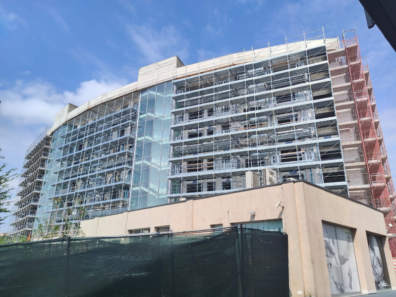 Moderno edificio a più piani con facciata in vetro, attualmente in costruzione, con impalcature e cielo azzurro sopra.