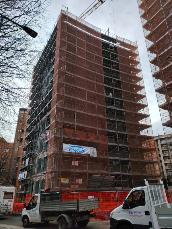 Un alto edificio in costruzione avvolto da impalcature rosse, con camion da lavoro parcheggiati al di sotto.