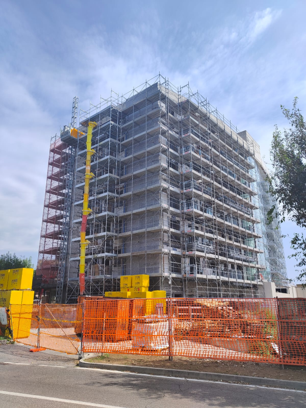 Edificio a più piani in costruzione, con impalcature, protetto da una recinzione di sicurezza arancione e sormontato dal braccio giallo di una gru.