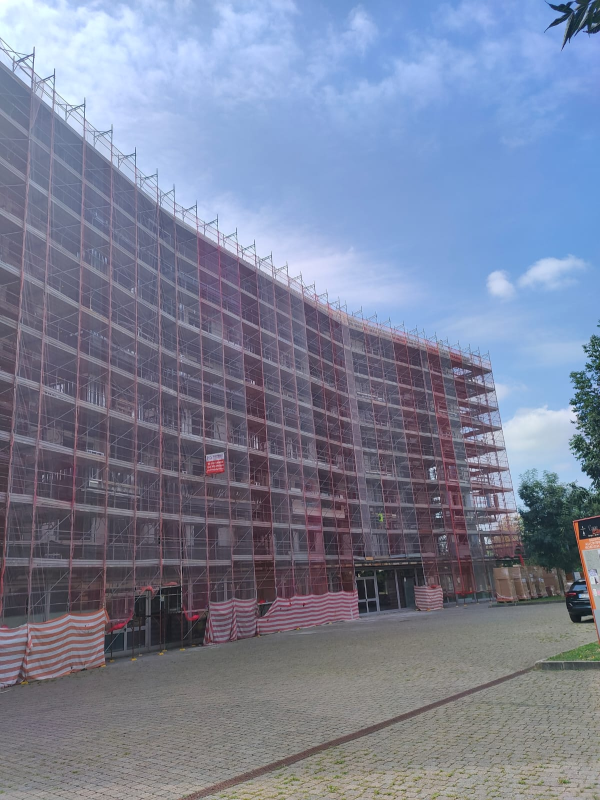 Edificio curvilineo avvolto da impalcature e reti rosse sotto un cielo azzurro, accanto a una piazza lastricata.
