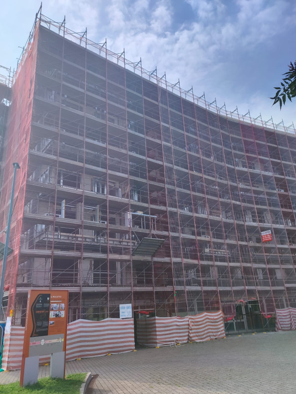 Impalcature avvolgono un edificio curvo in costruzione, nascosto dietro barriere arancioni e bianche.