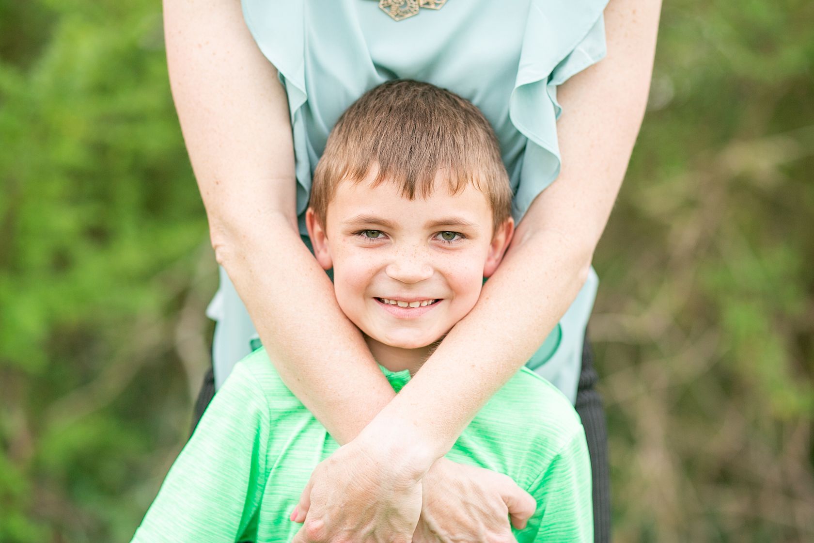 Smiling boy in a green shirt standing in front of an adult’s crossed arms outdoors