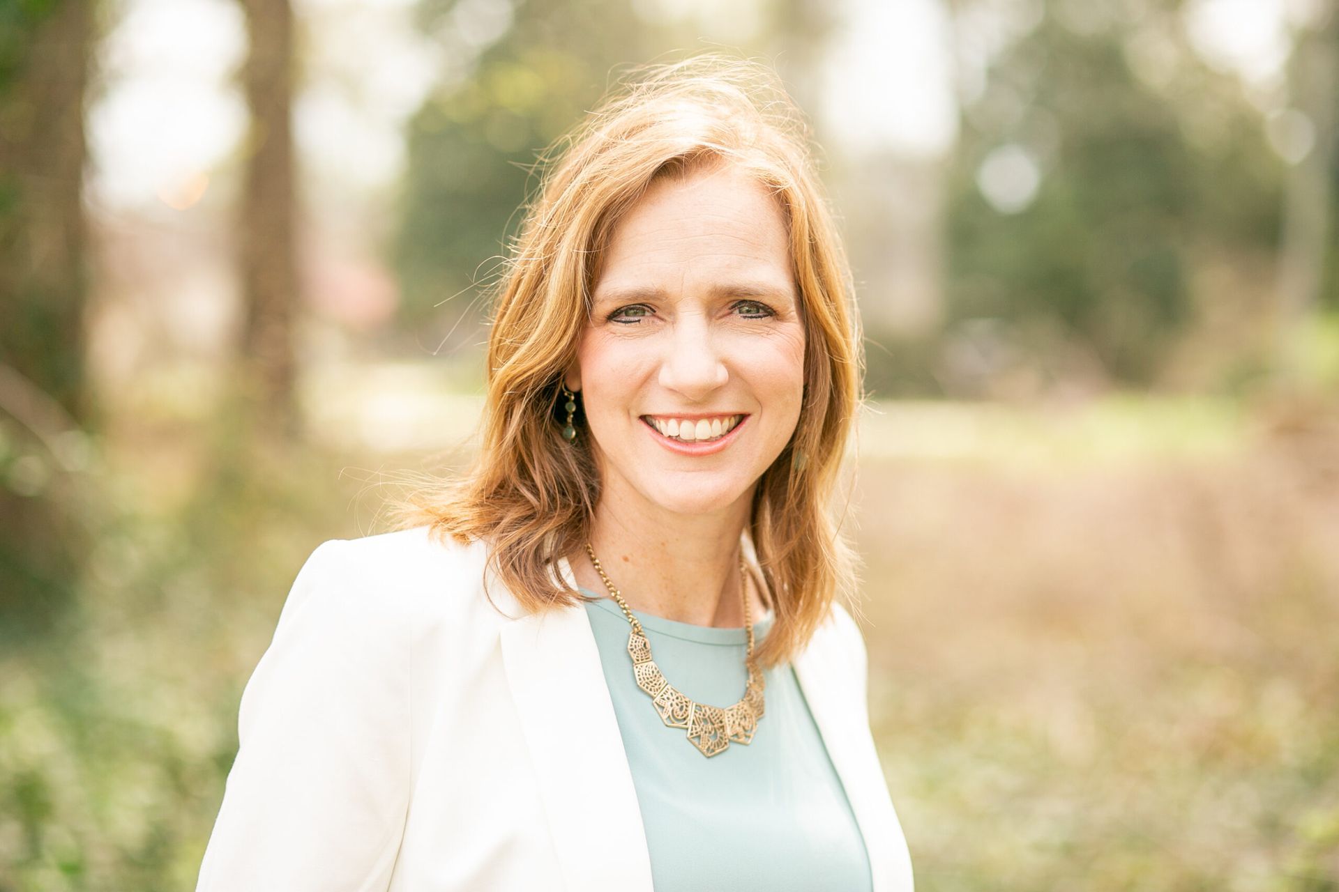 Smiling woman in a white blazer and teal top standing outdoors with a blurred garden background