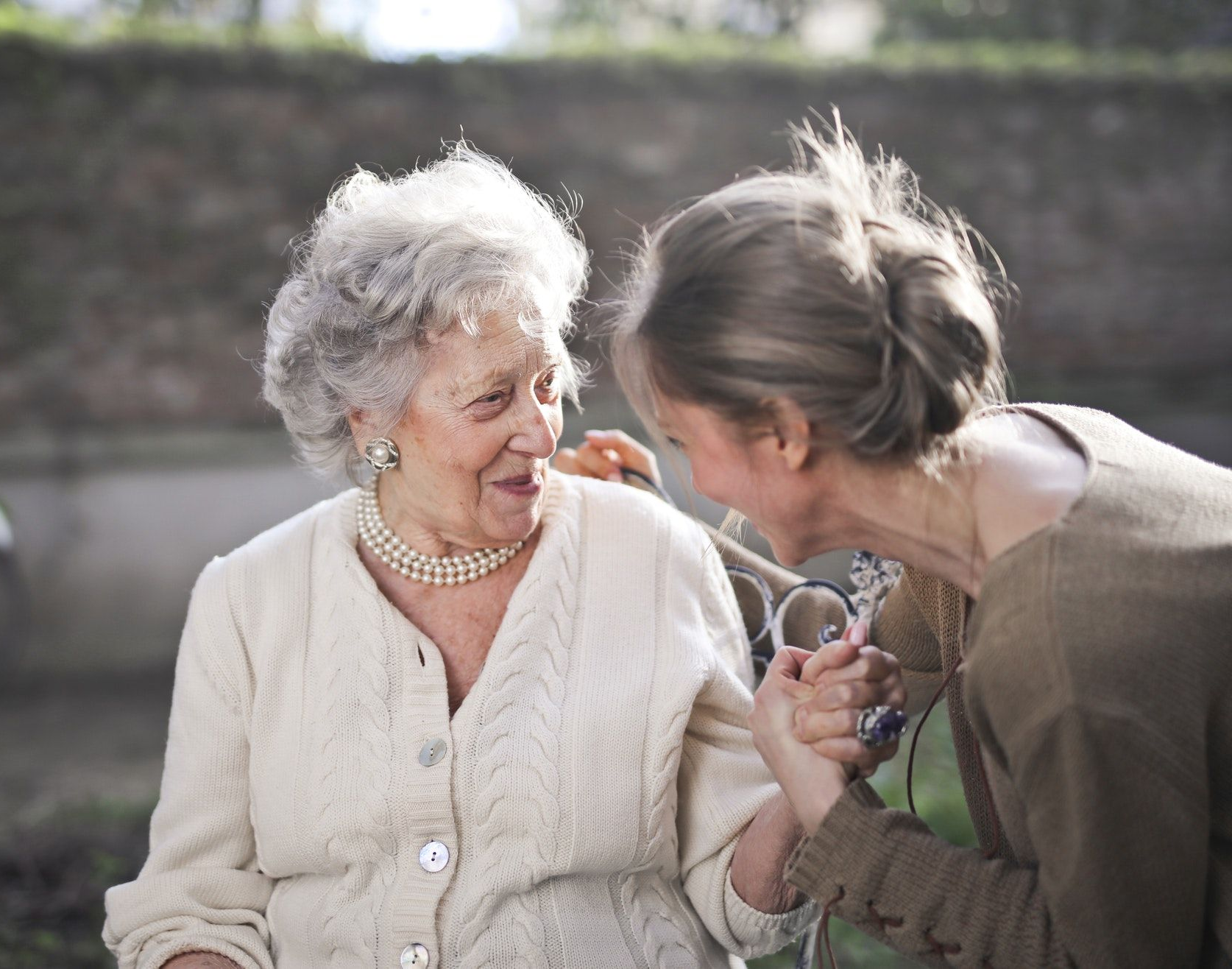 Two people smiling and talking outdoors, one holding the other's hand near a garden wall.