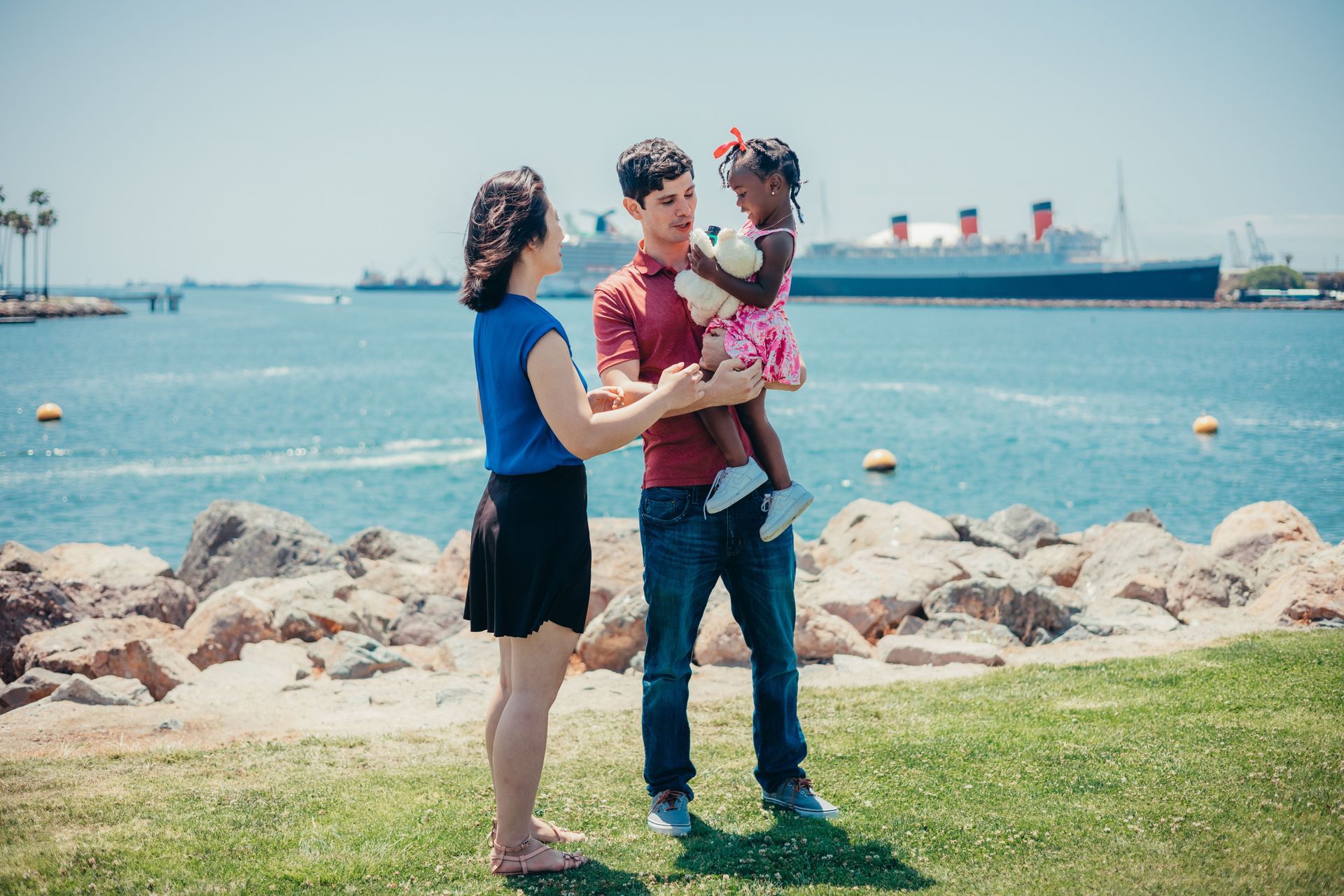 Family standing on a rocky shore, holding a child, with blue water and ships in the background
