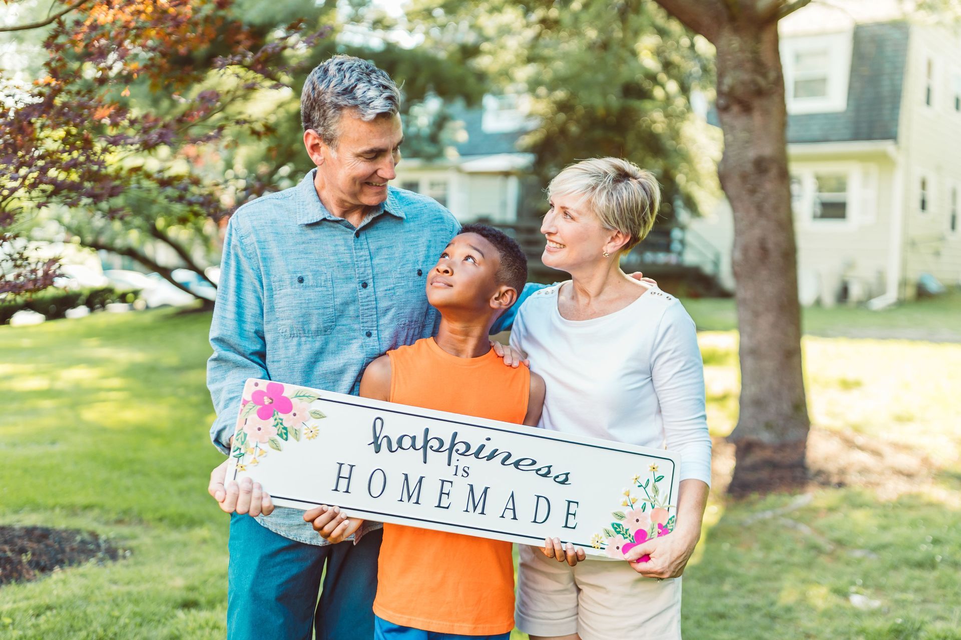Family holding a handmade sign in a sunny yard, smiling together with a house in the background