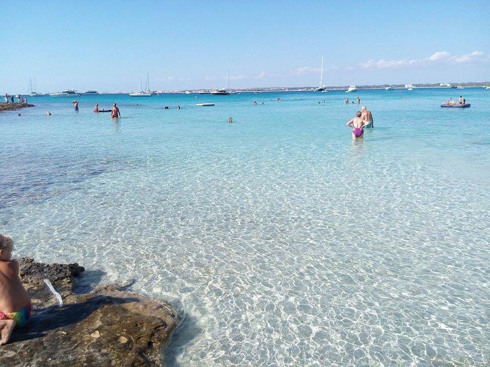Spiaggia dalle acque cristalline color turchese con bagnanti, barche in lontananza e una persona seduta sugli scogli in primo piano.