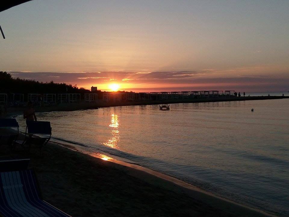 Tramonto su un lungomare tranquillo, con un riflesso luminoso e un lungo molo all'orizzonte.