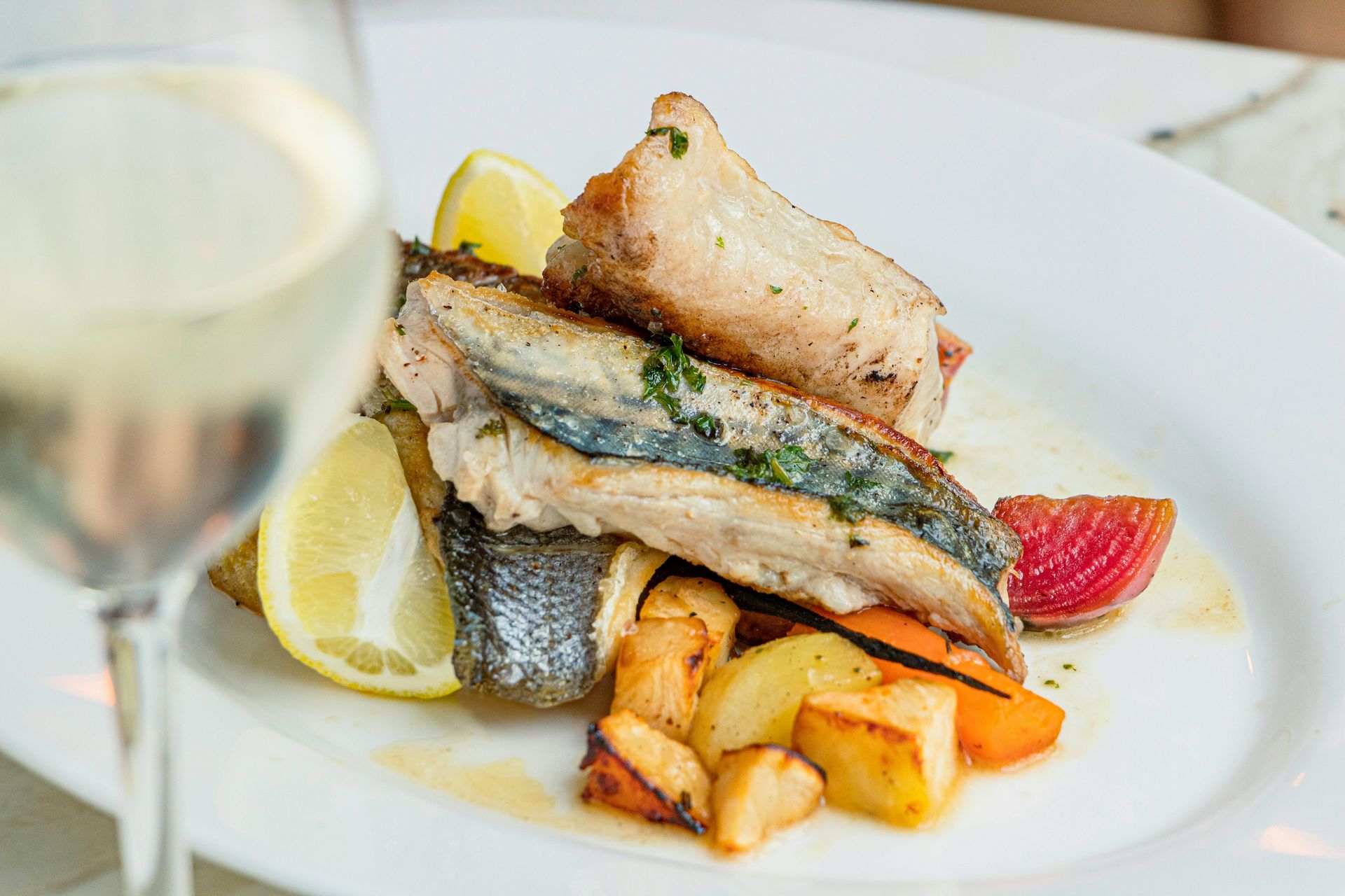 Pesce grigliato con spicchi di limone e patate arrosto su un tavolo bianco, con un calice di vino in primo piano.