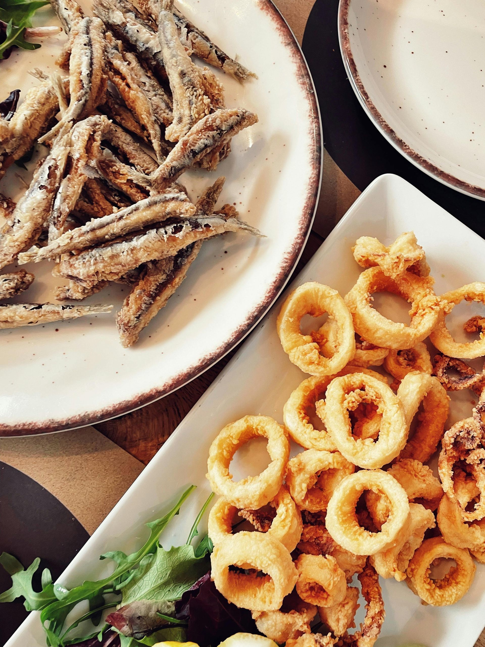 Piatti di pesciolini fritti e anelli di calamari dorati su un tavolo con verdure e salsa d'accompagnamento.