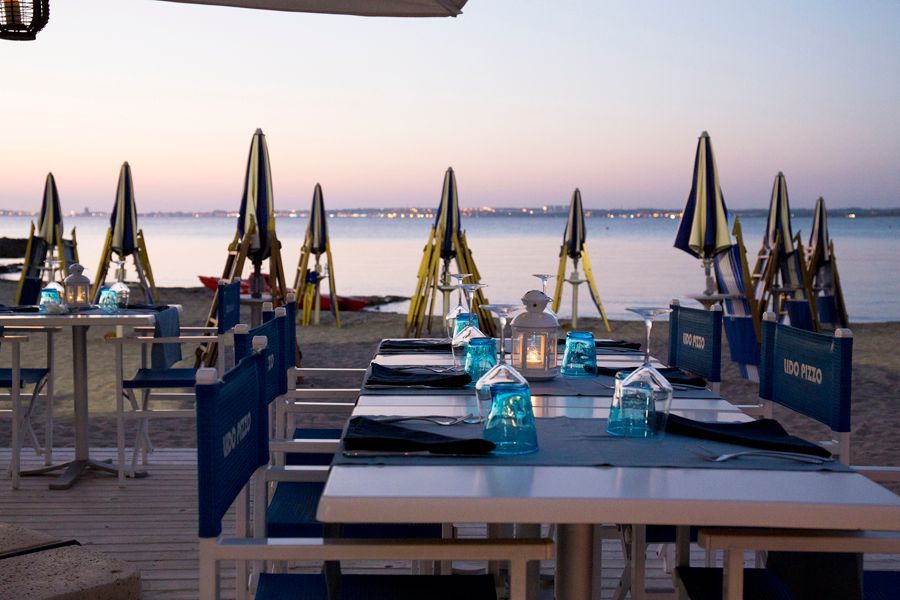 Tavoli apparecchiati per la cena al tramonto in un ristorante sulla spiaggia, con il mare e gli ombrelloni chiusi sullo sfondo.