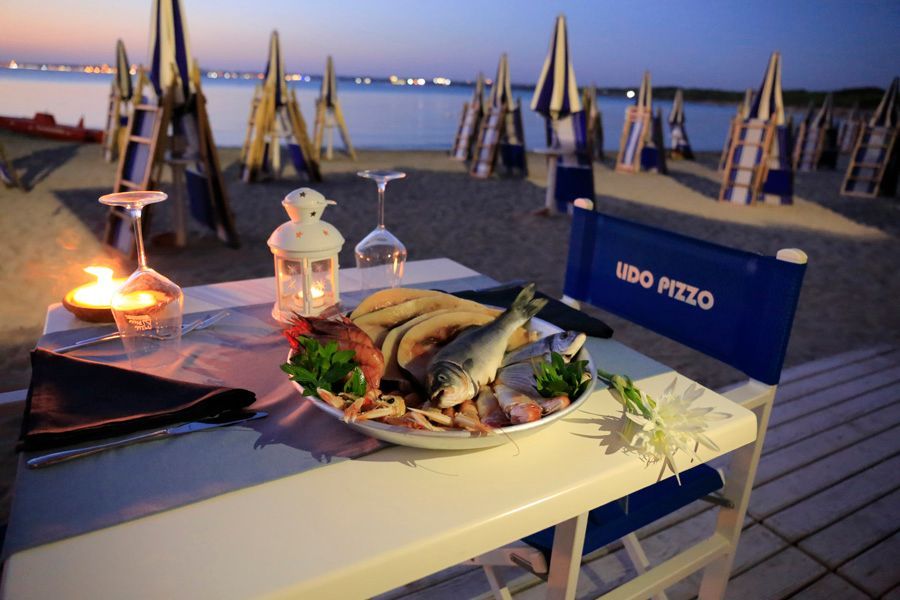 Cena a base di frutti di mare su un tavolo in riva al mare al tramonto, con l'oceano e gli ombrelloni di paglia sullo sfondo.
