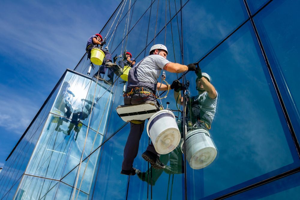 Two window washers in safety gear cleaning a tall blue glass building facade