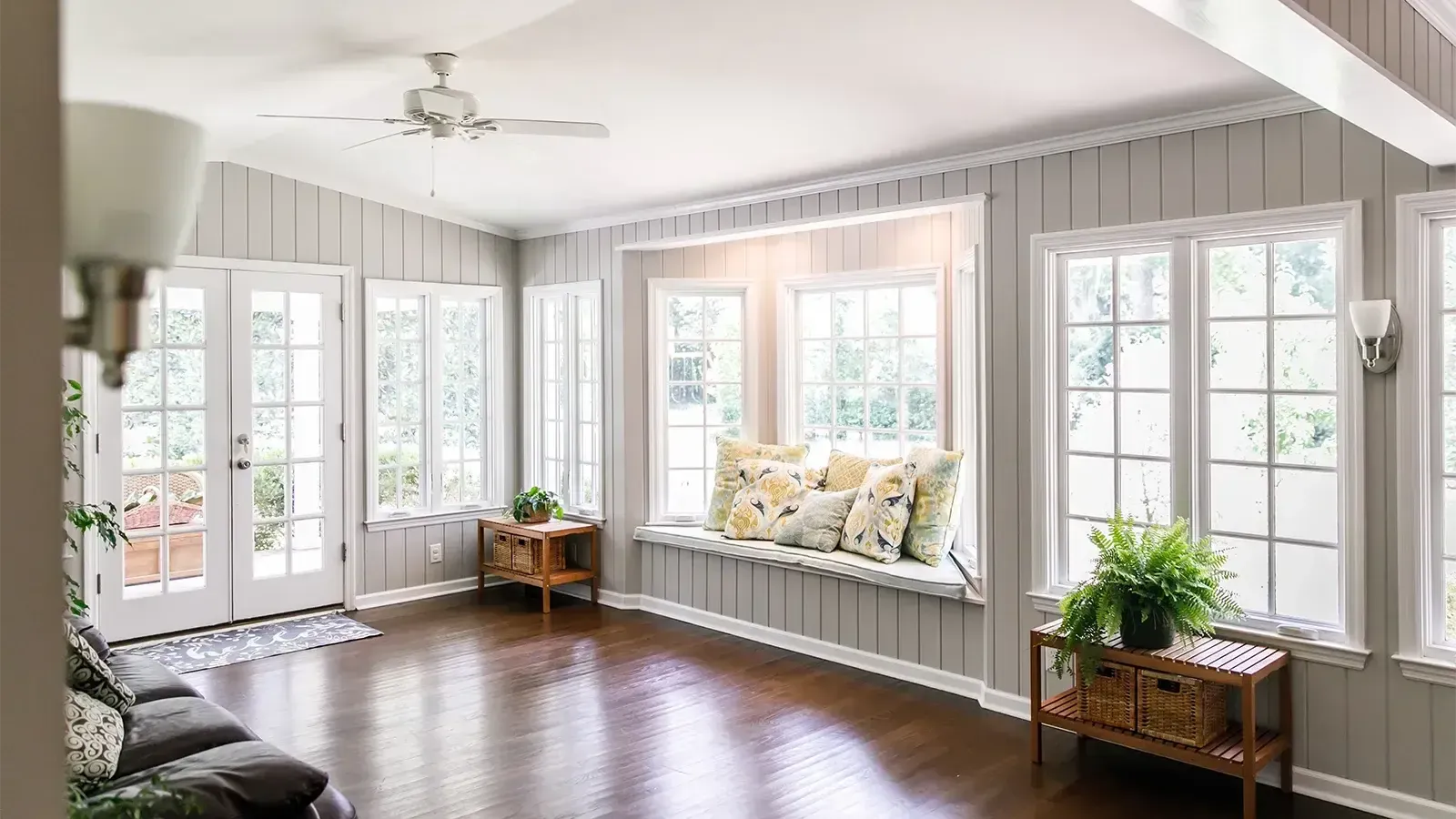Bright sunroom with many windows, dark wood floor, cushioned bench, and potted plants.