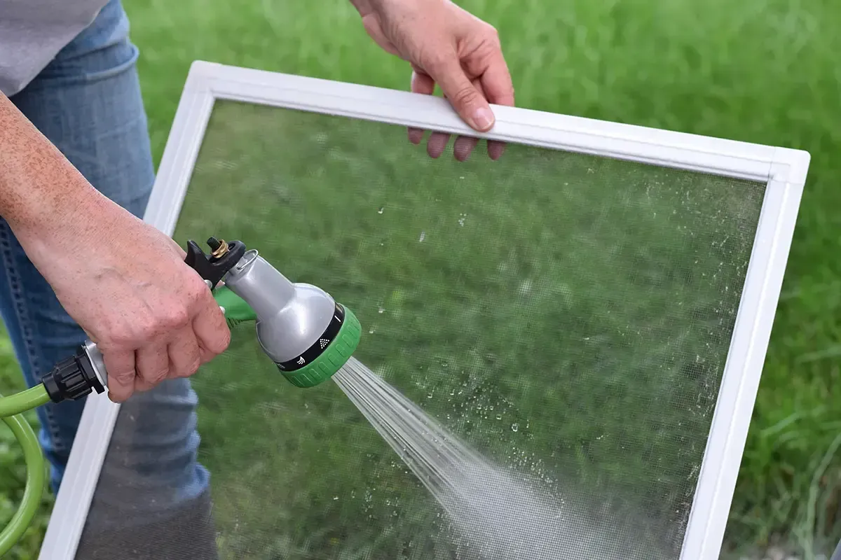 Person spraying a white window screen with a garden hose outdoors