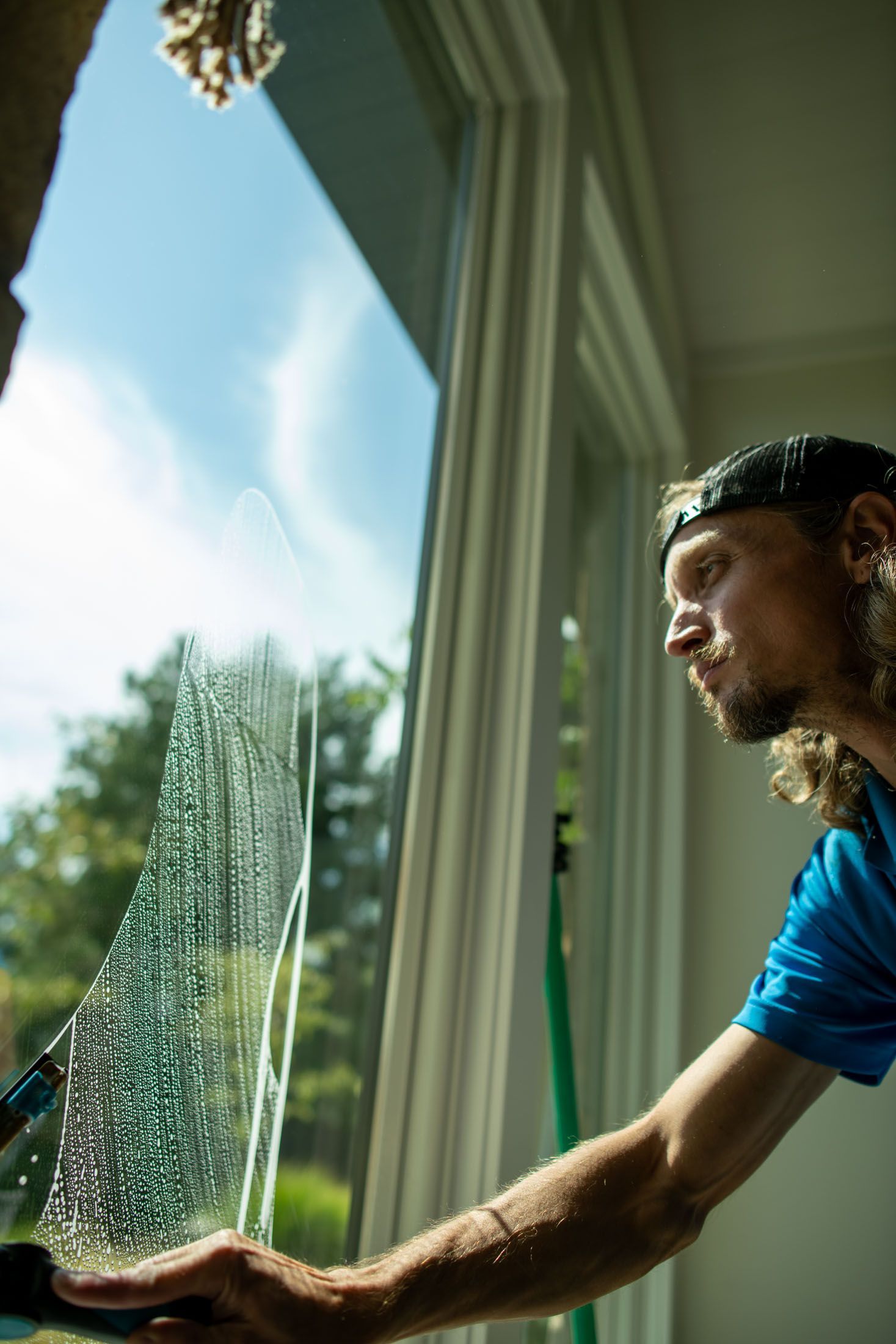 Window washer cleaning a window on a house exterior. Wearing blue shirt, working outside with mountain backdrop.