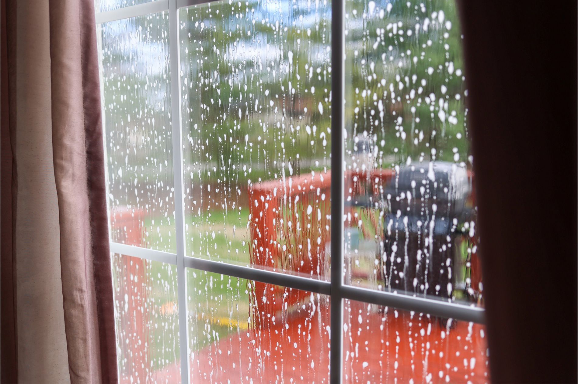 Rain-speckled window with curtains, overlooking a red patio and greenery outside.