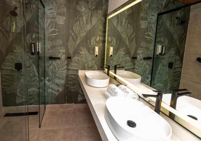 Baño moderno con papel tapiz de hojas verdes, dos lavabos blancos sobre una encimera larga y una ducha de cristal.