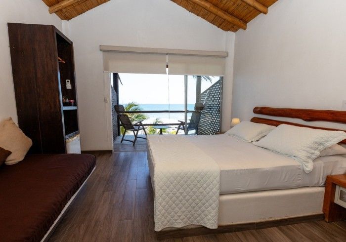 Dormitorio con vista al mar; paredes de madera, cama King, balcón con asientos y techo de paja.