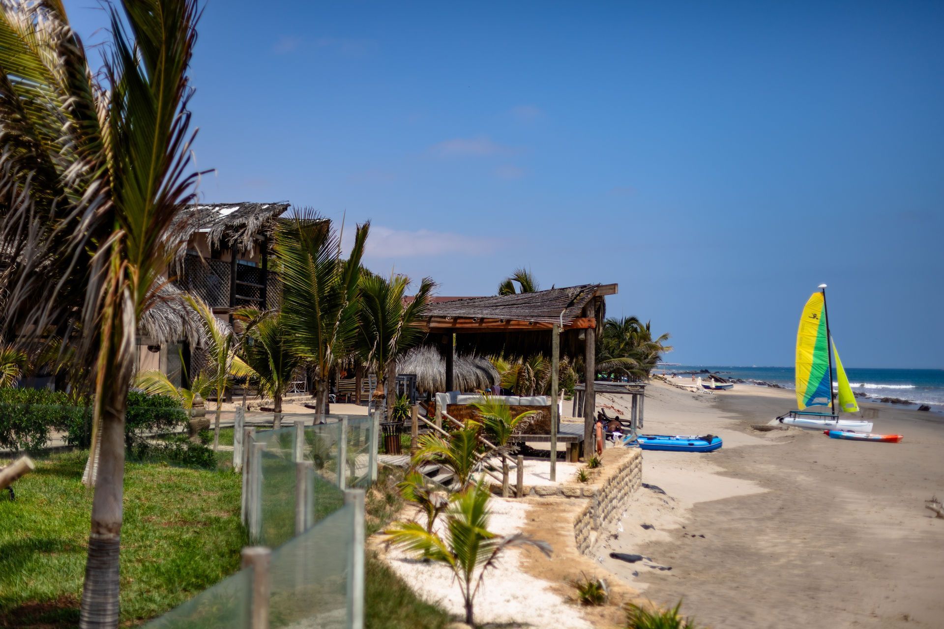 Escena de playa con palmeras, edificios con techo de paja y un velero en el agua bajo un cielo azul claro.