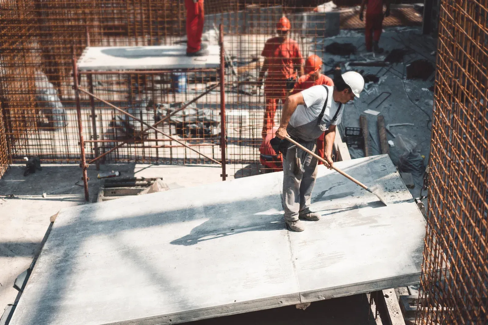 Construction worker leveling concrete on a slab at a building site, surrounded by rebar and other workers in red coveralls.
