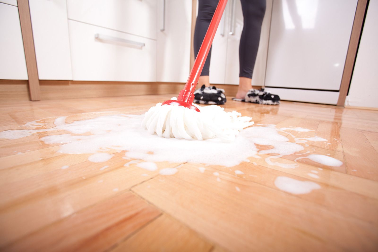 Person mopping a wooden floor with a red-handled mop and soapy water in a kitchen.