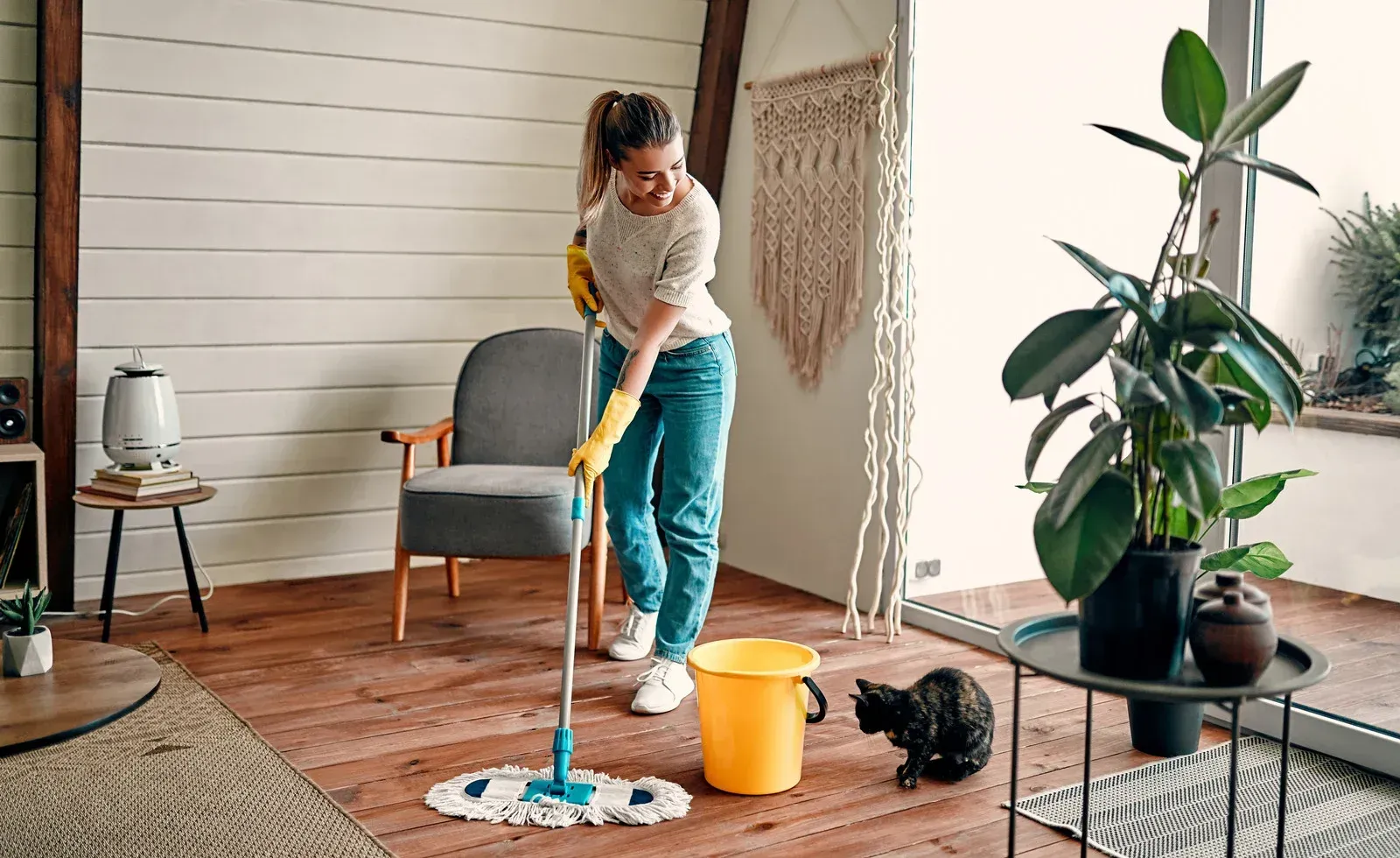 Woman mopping a wooden floor in a living room, wearing gloves. A black cat watches nearby.