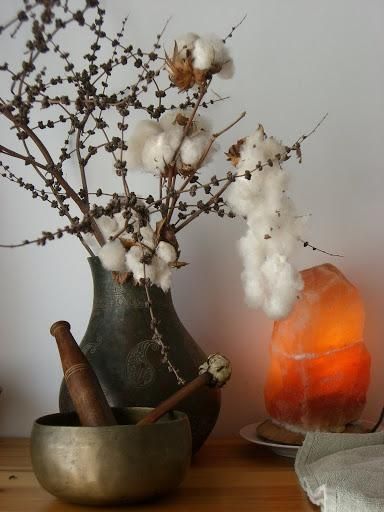 Cotton stems in a dark vase beside a bronze bowl and glowing orange salt lamp on a white surface