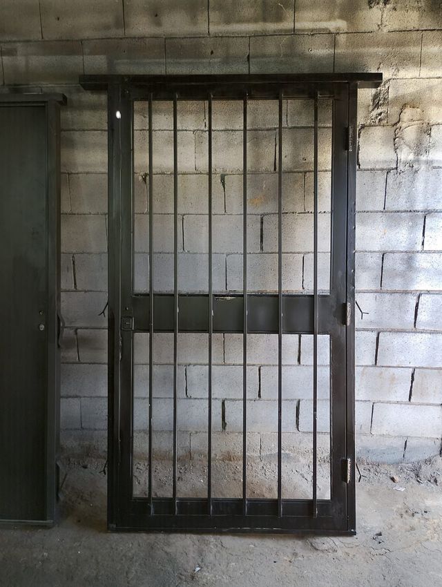 Una verja metálica en un muro de bloques de hormigón, con una puerta oscura a la izquierda.