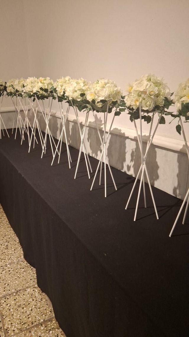 Arreglos florales blancos sobre altos pedestales alineados sobre una mesa cubierta con un mantel