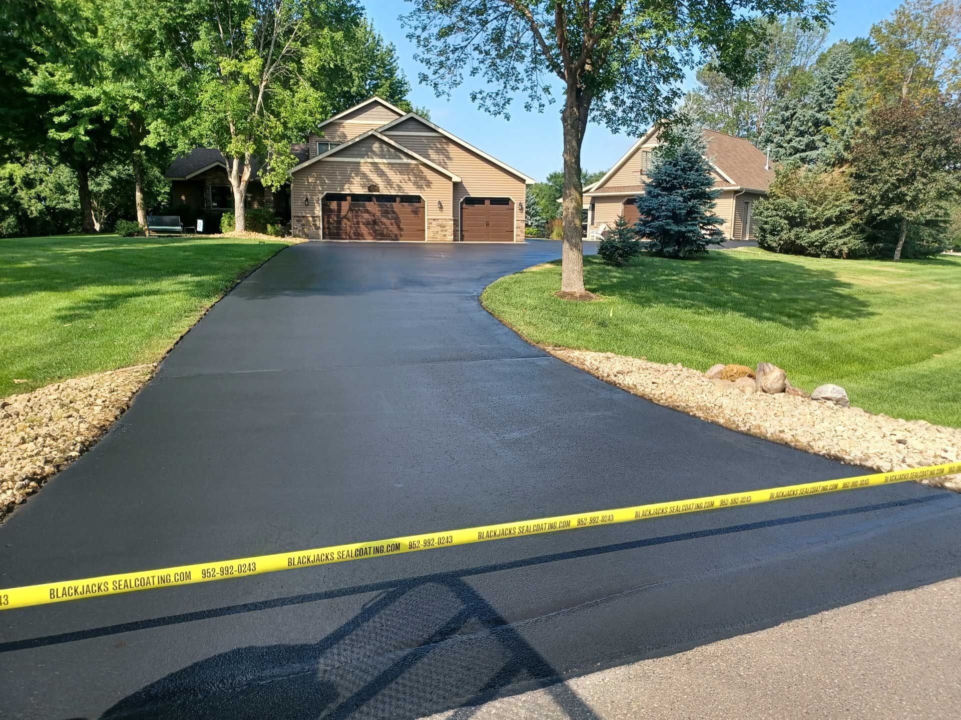 Freshly paved black driveway leading to a suburban garage, blocked by yellow caution tape under bright trees