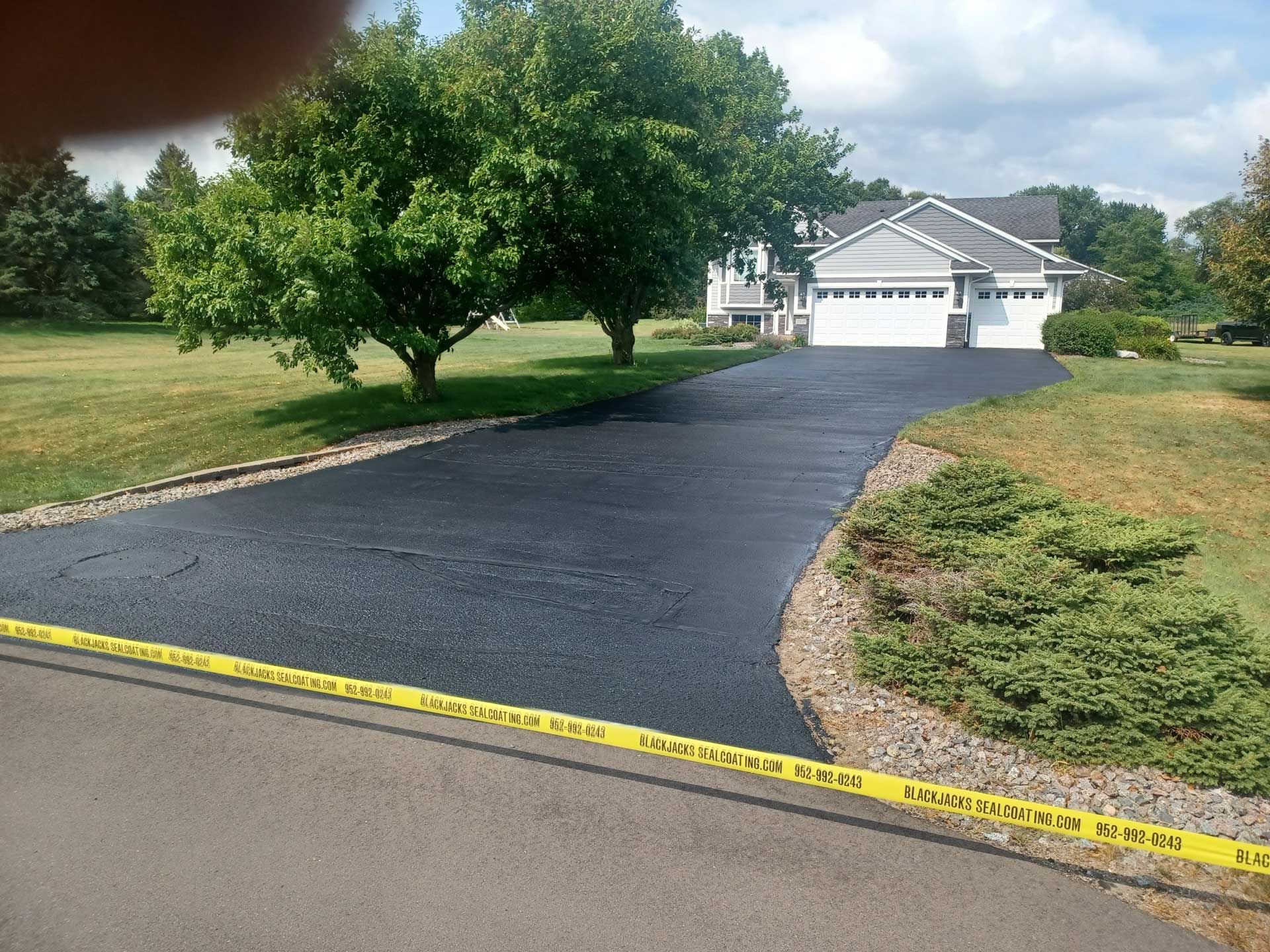 Freshly paved black driveway leading to a white garage, bordered by yellow caution tape and trees