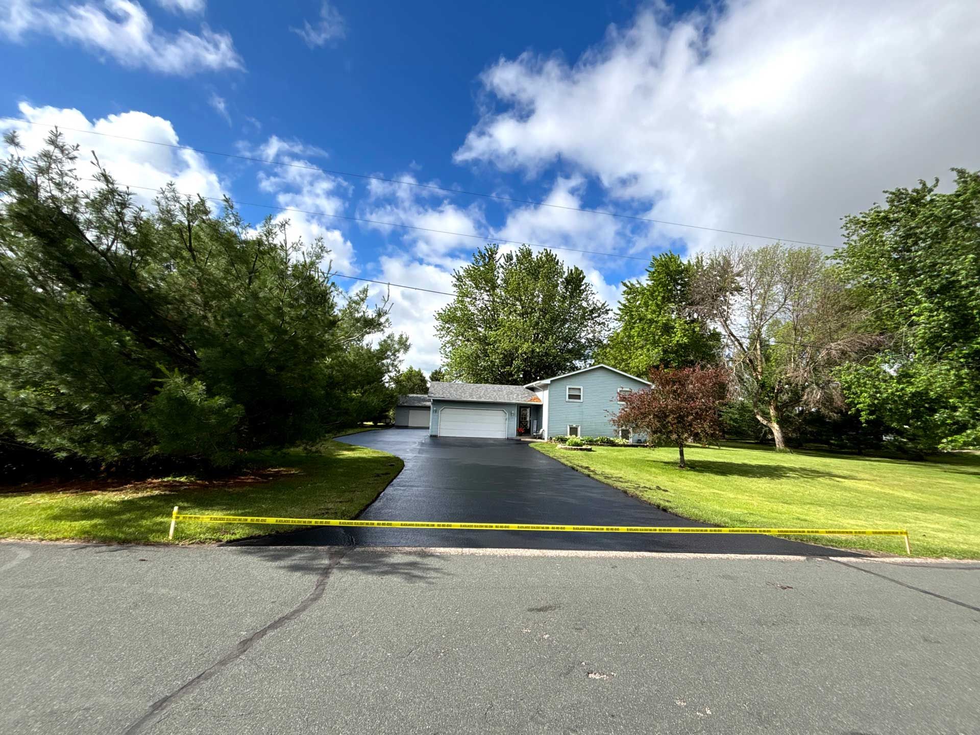 Residential driveway leading to a white house, lined with green trees and lawn under a partly cloudy sky