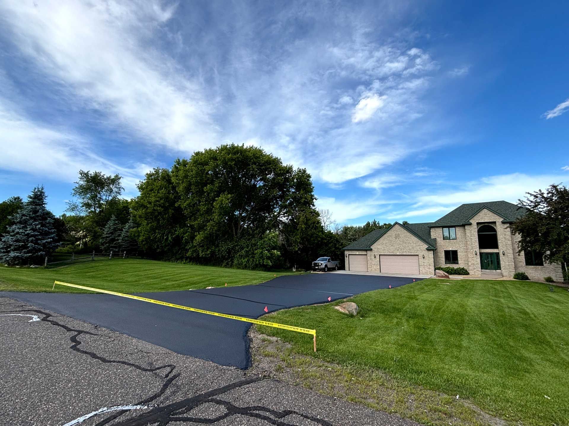 Driveway and stone house under a blue, cloudy sky, with trees and green lawn