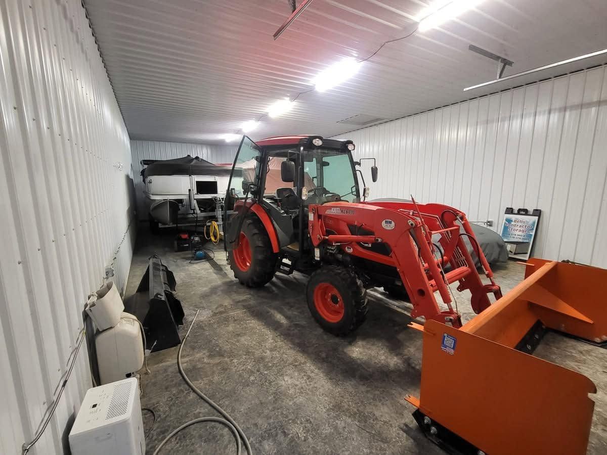 Red tractor with front loader in a white storage garage, with orange equipment in the foreground.