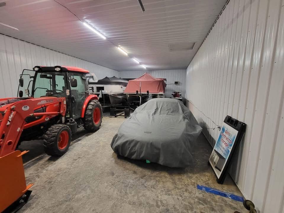 Covered car in a white-walled garage beside a red tractor and other equipment.