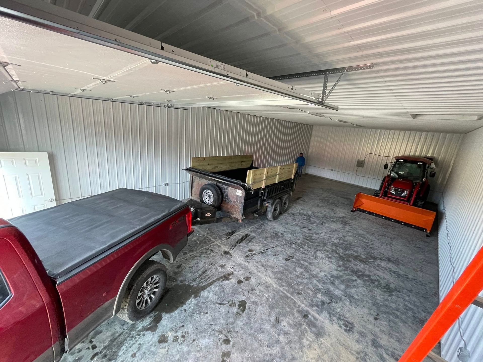 Covered garage with a red pickup, a beige utility trailer, and an orange lawn tractor on a concrete floor